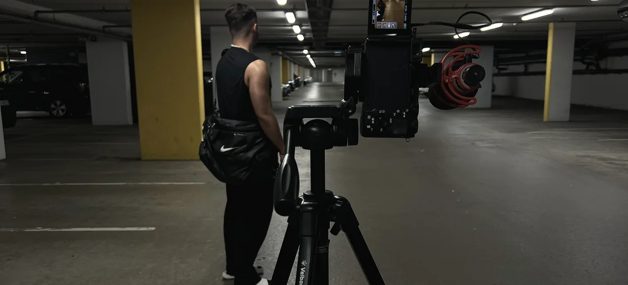 A camera with a red microphone on a tripod in an underground car park, a crew member beside it